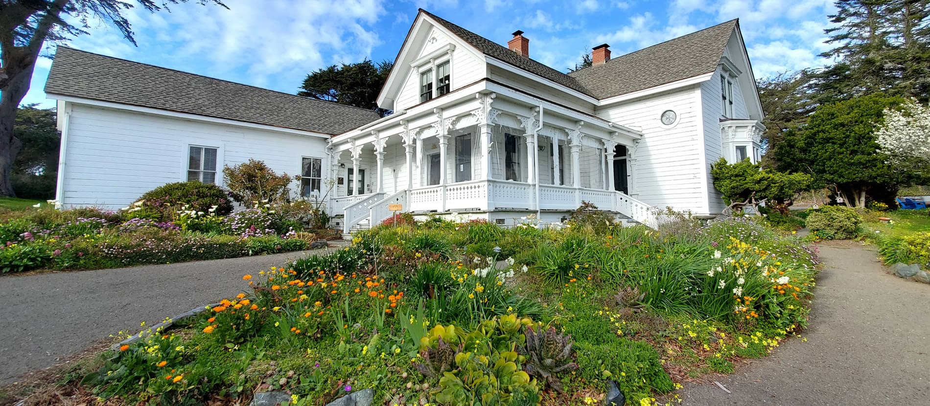A picturesque white Victorian house surrounded by vibrant flowers under a blue sky.