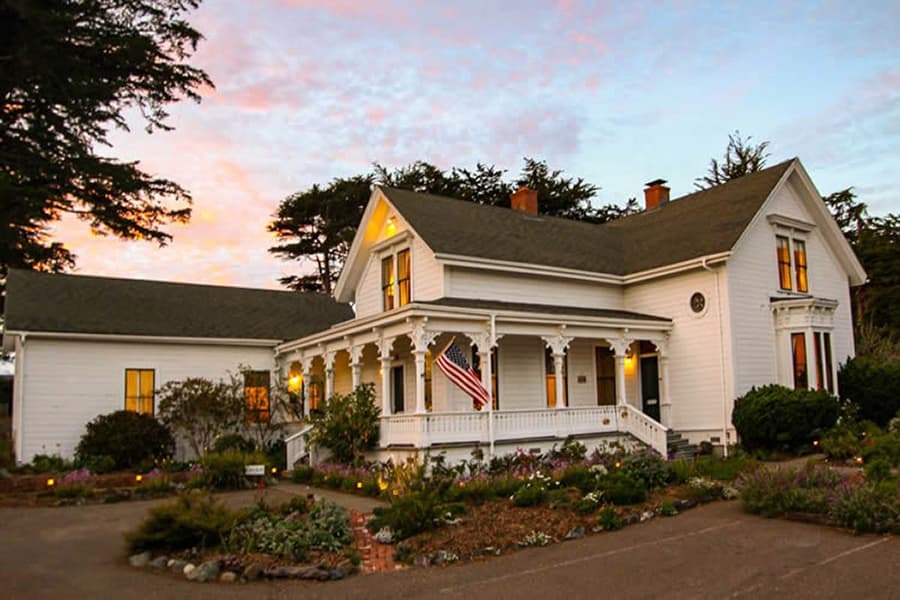 A charming white house with a porch and an American flag, surrounded by lush gardens at sunset.
