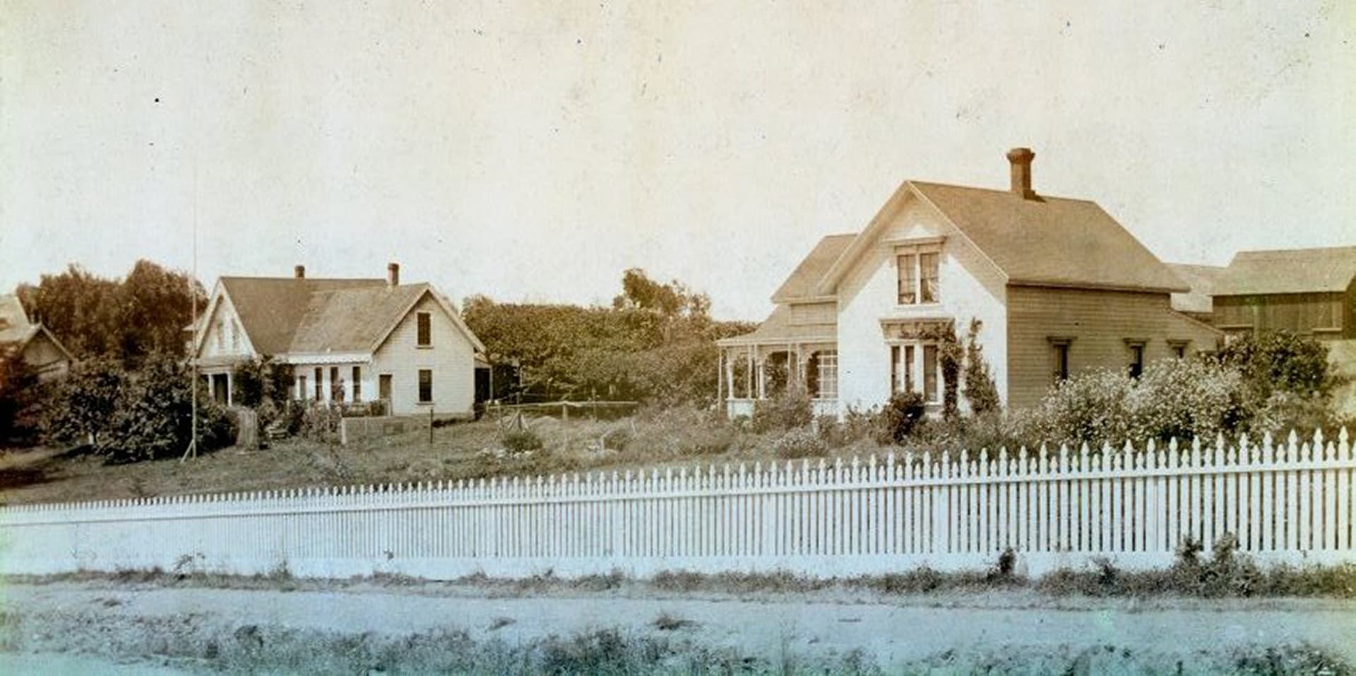 Two historical houses surrounded by greenery behind a white picket fence.
