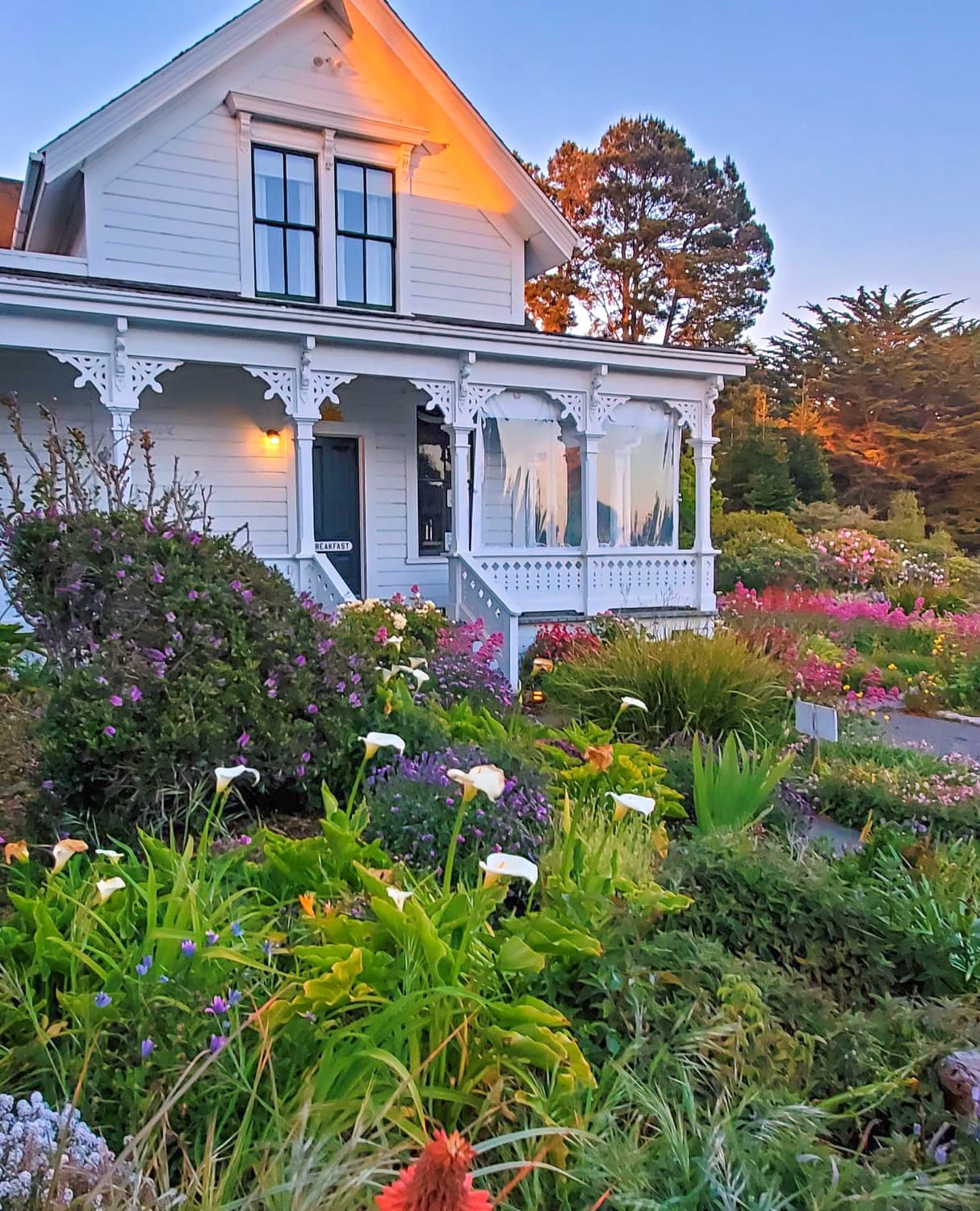 A charming white house with a decorative porch is surrounded by a vibrant garden of flowers.