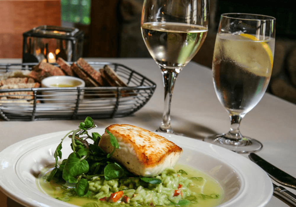 A seared fish fillet on a bed of greens, accompanied by white wine and a glass of water, with a basket of bread in the background.