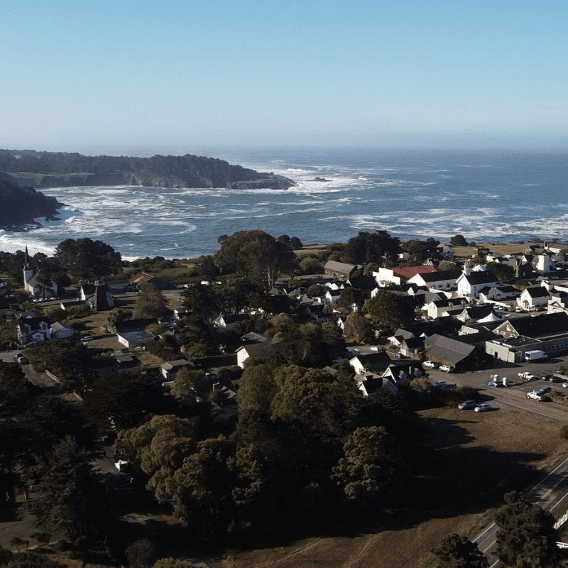 Aerial view of a coastal town with ocean waves crashing nearby and green hills in the background.