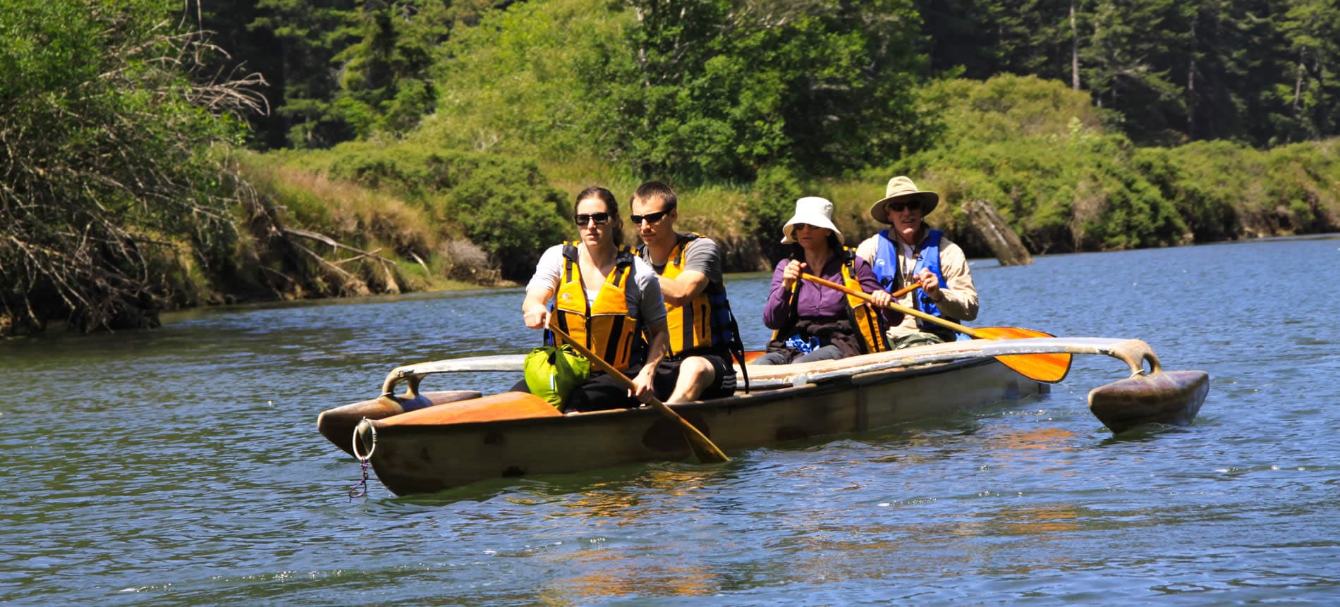 A group of four people paddles in a canoe on a calm river surrounded by greenery.