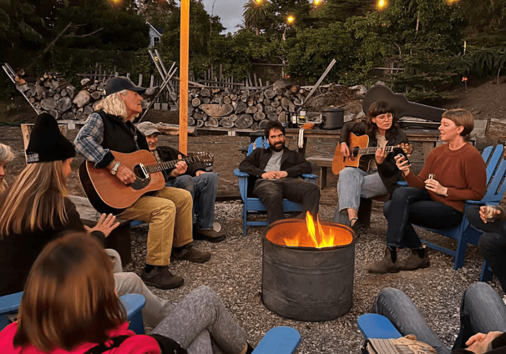 A group of people gather around a fire, playing guitars and sharing music in a cozy outdoor setting.