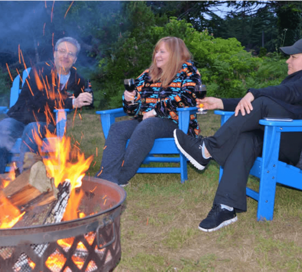 Three people enjoy wine by a fire pit on a grassy area, seated in blue chairs.