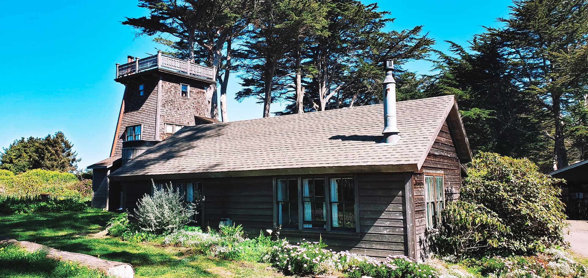 A rustic wooden house with a tower, surrounded by lush greenery and tall trees under a clear blue sky.
