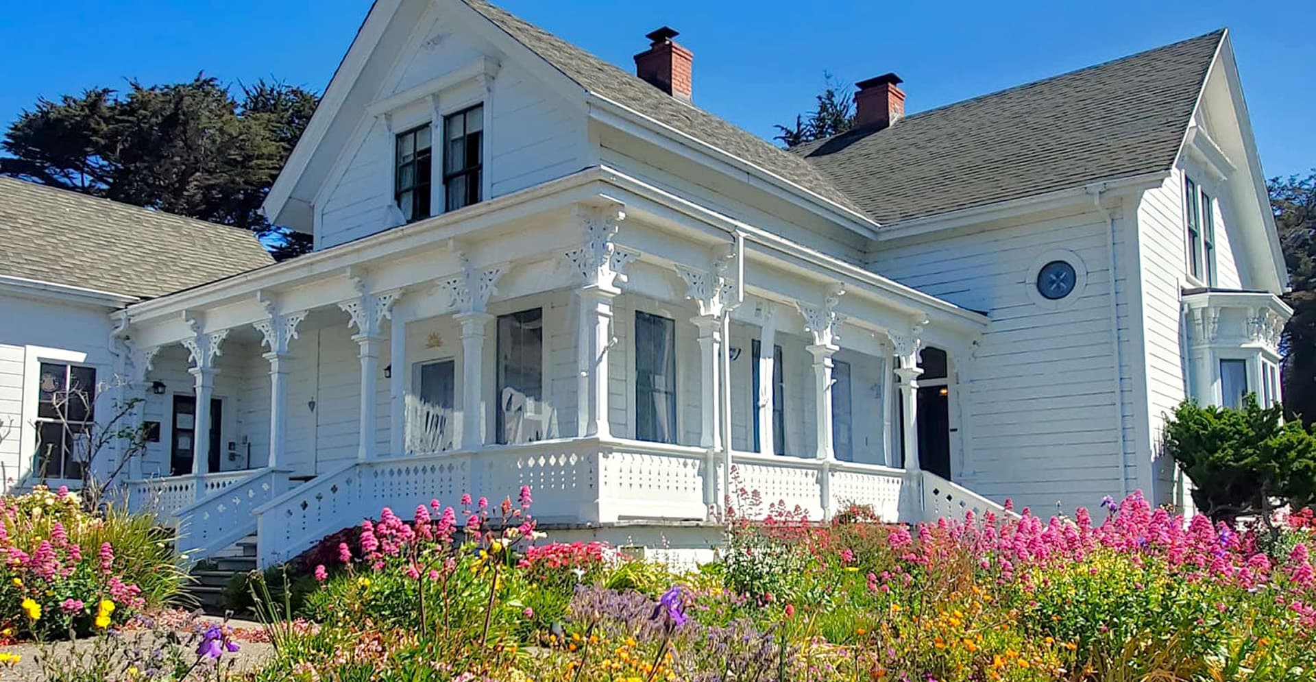 A charming white Victorian house surrounded by vibrant flowers under a clear blue sky.
