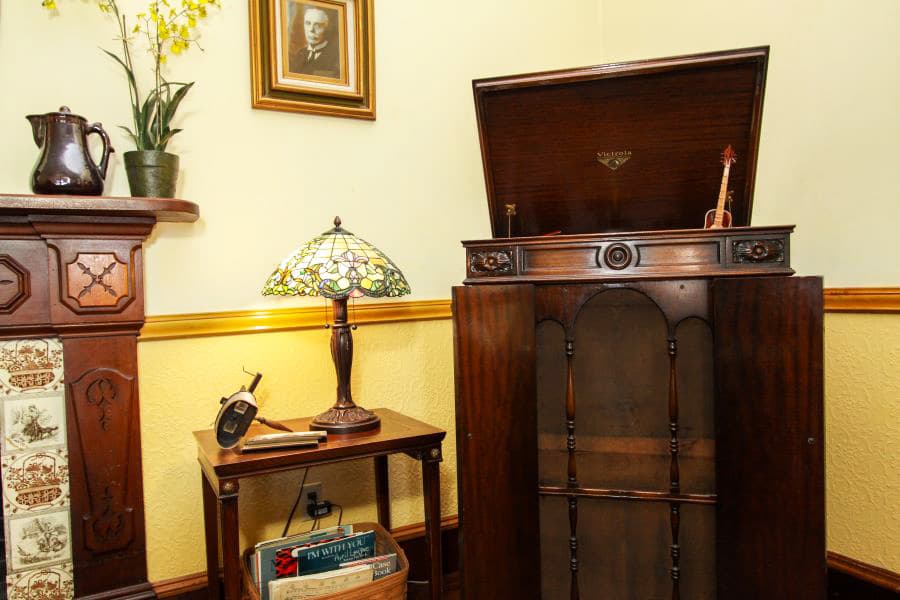 A vintage wooden phonograph next to a table lamp and a bookshelf in a warmly decorated room.