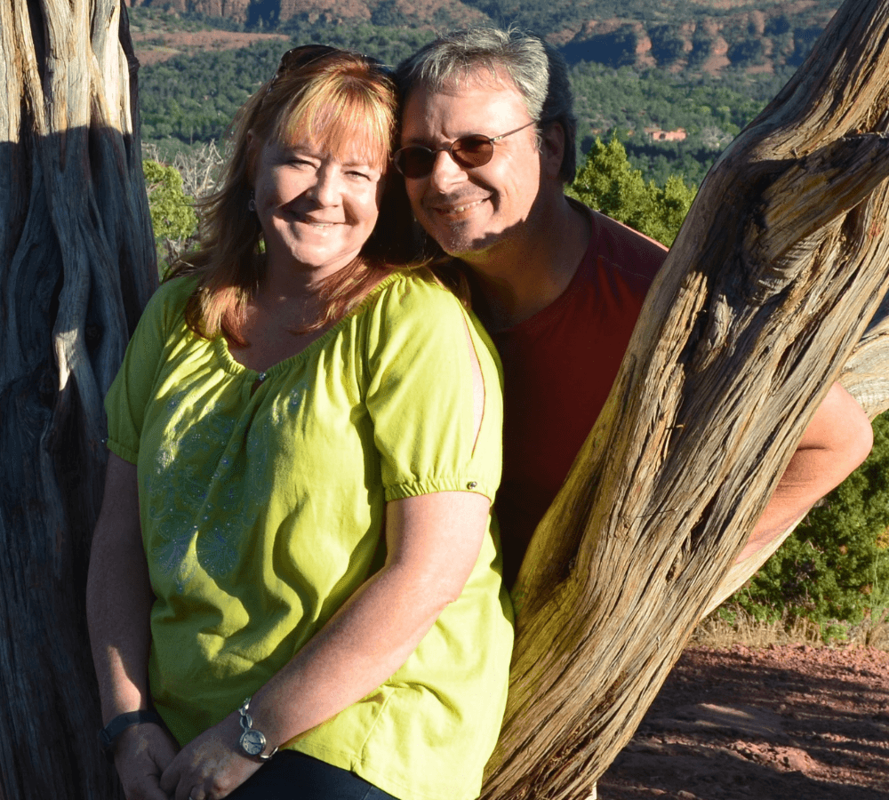 A smiling couple poses together outdoors against a backdrop of trees and mountains.