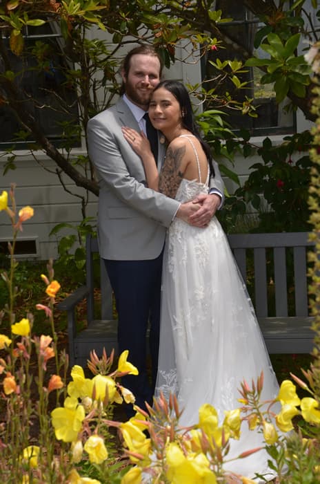 A couple embraces on a bench surrounded by flowers, with the woman wearing a wedding dress and the man in a light gray suit.