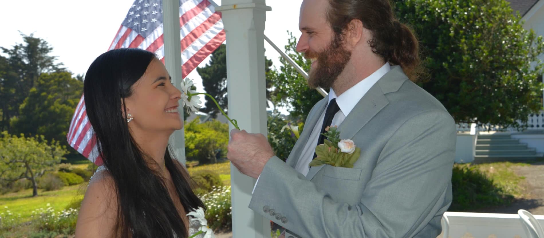 A smiling couple gazes at each other during a wedding ceremony, with an American flag in the background.