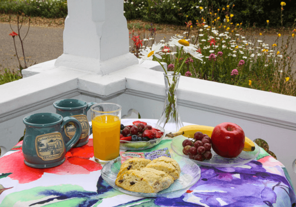 A colorful table setting features two mugs, a glass of orange juice, assorted fruits, and a plate of scones, with a garden in the background.