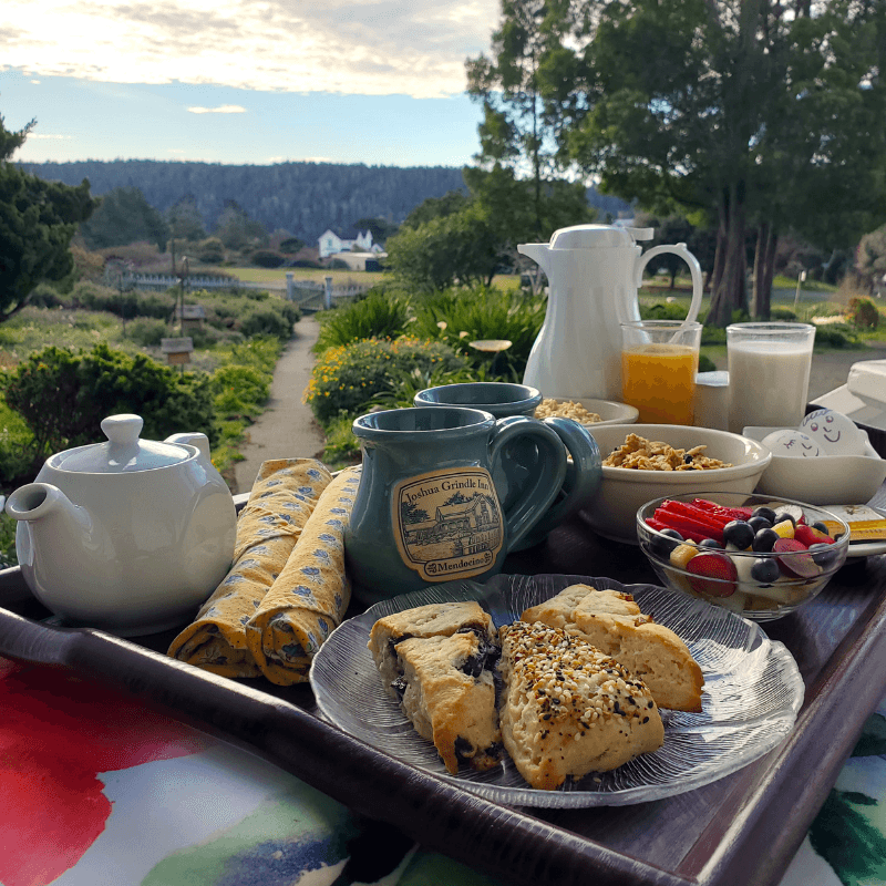 A beautifully arranged breakfast tray featuring tea, coffee, pastries, yogurt, fruit, and a picturesque garden view.