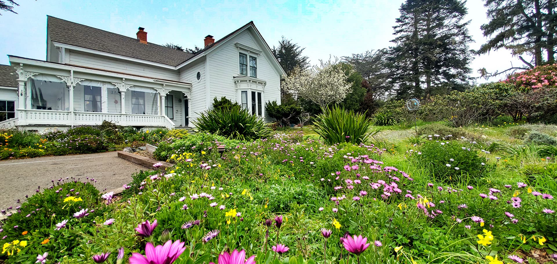 A white Victorian house surrounded by vibrant flowers and greenery.